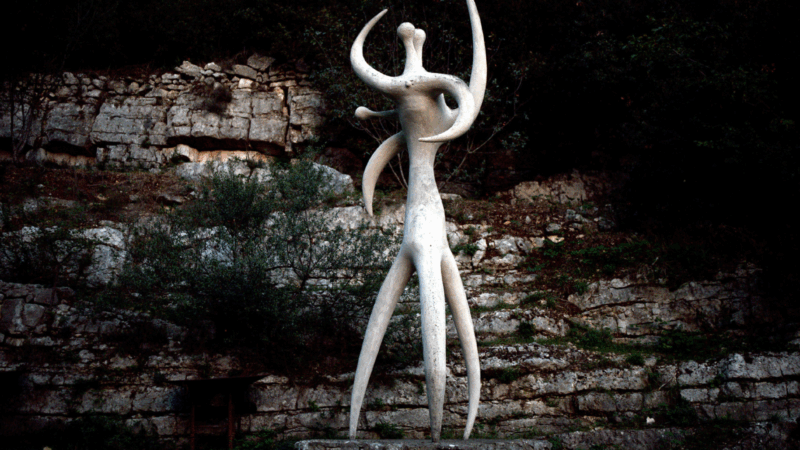 Uma escultura abstrata e orgânica de Robert Roussil, de cor bronze ou marrom, com formas longas e sinuosas que lembram um animal deitado ou raízes. A escultura está sobre um gramado, com vegetação e um paredão rochoso ao fundo, onde outra escultura branca é vista à distância.