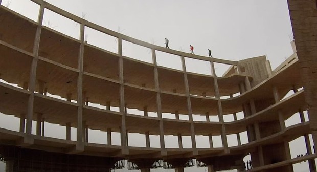 Cena do filme Yalla Parkour Na imagem, três garotos caminham no topo de uma estrutura de um aeroporto abandonado. O céu está nublado e não dá para identificar os garotos, pois a câmera está bem afastada. O edifício não possui portas ou janelas, apenas sua estrutura crua. 