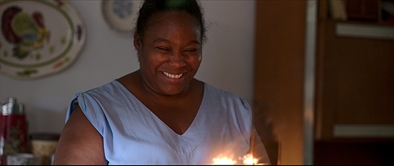 Cena do filme Criadas. Sandra, uma mulher de pele negra e cabelos crespos presos, sorri com alegria olhando para velas acesas em cima de um bolo, que não aparece na imagem. Ela veste uma blusa azul-clara. No fundo, há uma decoração composta por pratos de porcelana na parede.