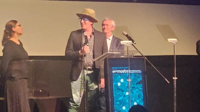 Foto em ângulo baixo de um homem discursando em um palco durante o evento. Ele é o foco central, usa um chapéu bege, óculos de grau com armação azul vibrante, blazer escuro sobre uma camisa estampada e calça jeans. Ele segura o microfone com a mão direita e sorri. À sua frente, há um púlpito de acrílico exibindo o cartaz azul da 49ª Mostra Internacional de Cinema de São Paulo. Ao fundo, levemente desfocado à direita, um homem de cabelos grisalhos e terno observa sorrindo, e à esquerda, uma mulher de preto observa o orador.