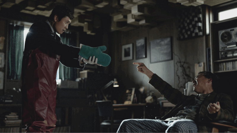 Dentro de uma sala com luz difusa, um homem de avental vermelho e luvas verdes estende as mãos na direção de outro homem, que está sentado e reage com surpresa, apontando o dedo para ele. O ambiente é repleto de móveis escuros, quadros e equipamentos de som, sugerindo um momento de confronto contido.