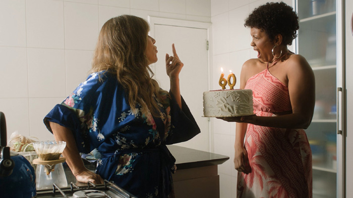 Cena do filme Sexa. Em uma cozinha iluminada, Bárbara, de roupão azul florido, gesticula com o dedo médio levantado enquanto conversa com Cristina. Ela usa um vestido rosado estampado e a olha para a amiga com uma expressão indignada enquanto segura um bolo branco com velas douradas formando o número 60.