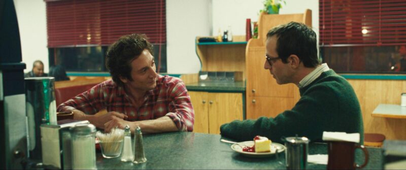 Dois homens brancos sentados em uma mesa de um restaurante, homem da esquerda de camisa de flanela vermelha e homem da esquerda de suéter verde os dois estão conversando