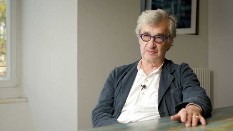Cena do ensaio videográfico Filmar ou Morrer. O cineasta Wim Wenders está sentado diante de uma mesa, olhando com expressão séria para alguém atrás da câmera. Ele veste um casaco escuro e camisa branca, e seus óculos arredondados chamam atenção. Ao fundo, uma parede clara com um quadro e uma janela iluminam suavemente o ambiente.