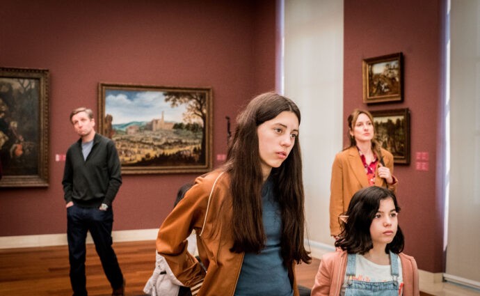 Cena do filme Maturidade. Na imagem, da esquerda para a direita, é possível ver quatro pessoas: um homem branco vestindo calças pretas e uma jaqueta preta; uma jovem adolescente de pele branca e cabelo longo castanho, vestindo uma blusa azul e uma jaqueta marrom; uma criança de pele branca e cabelos castanhos curtos, vestindo uma camiseta branca, um macacão jeans e uma jaqueta rosa; e uma mulher de pele branca, vestindo uma blusa colorida e um casaco marrom. Eles estão em um museu, onde é possível ver pinturas emolduradas em uma parede marrom-avermelhada. O chão do museu é de madeira e a luz predominante na imagem é clara