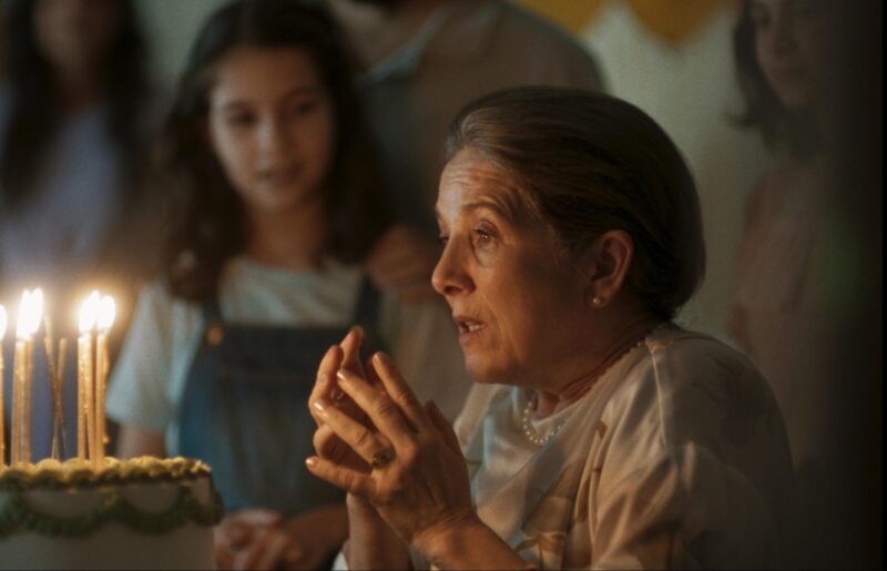 Cena do filme Querido Trópico. Uma mulher mais velha, de cabelos presos e grisalho está usando brincos e um colar de pérola e um vestido de tons claros. Ela está diante de um bolo com velas acesas e está com as mãos erguidas como se estivesse cantando "Parabéns". Ao fundo, há uma menina com cabelos escuros que observa a cena, levemente desfocada. 