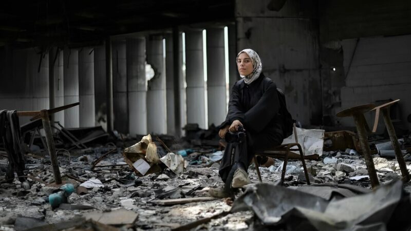 Cena do filme Guarde o Coração na Palma da Mão e Caminhe.Na imagem, Fatima Hassouna está no canto direito, sentada em uma cadeira. O ambiente é o interior de um prédio, que foi destruído por bombardeios. Há vários escombros no chão. Hassouna é uma mulher palestina, na faixa dos 25 anos, ela usa uma roupa inteira preta, mochila preta nas costas e está sentada com as pernas cruzadas. Nos pés, usa um tênis branco. Suas mãos estão apoiadas no joelho e em uma delas há uma câmera. Na cabeça, usa um lenço de sua cultura chamado de hijab, na cor branca e estampado. Hassouna olha para a frente, encarando a câmera. 