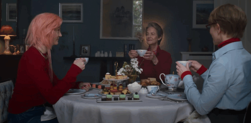 Cena do filme Pai Mãe Irmã Irmão. Três mulheres sentadas ao redor de uma mesa redonda posta para um chá da tarde elegante. À esquerda, uma mulher de perfil com cabelo rosa e suéter vermelho segura uma xícara. Ao centro, uma mulher mais velha com cabelos curtos e grisalhos sorri levemente enquanto ergue sua xícara. À direita, uma terceira mulher de camisa azul clara está de costas para o observador. A mesa está coberta por uma toalha branca e repleta de louças de porcelana florida, macarons e doces finos. O ambiente tem paredes azuis escuras e quadros ao fundo.