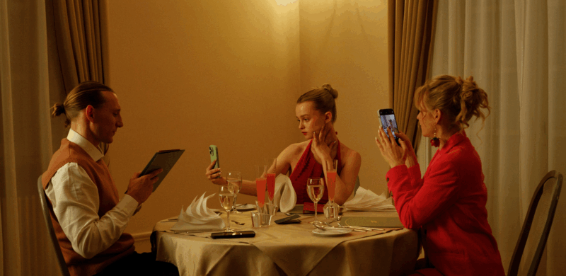 Cena do filme Babystar: À esquerda, um homem de cabelo longo preso em um coque olha para uma tela. No centro, Luca, uma mulher branca e loira, de vestido vermelho amarrado ao pescoço, tira uma selfie com o celular, enquanto sua mãe, à direita, também branca e loira em um conjunto rosa, fotografa a cena com o celular. Todos estão sentados em uma mesa de jantar elegante, iluminada por luz amarela suave, com taças, guardanapos dobrados e cortinas de tons claros.