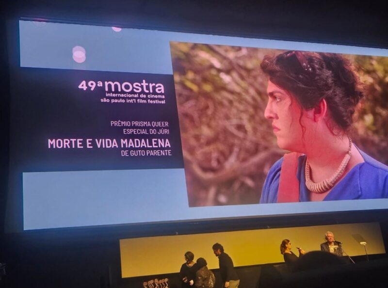 Fotografia da cerimônia de encerramento da 49ª Mostra Internacional de Cinema em São PauloNa imagem, em uma tela projetada, há o encarte da 49ª Mostra Internacional de Cinema em São Paulo, que anuncia o filme Morte e Vida Madalena como o vencedor do Prêmio Prisma Queer Especial do Júri. No canto esquerdo, em um retângulo preto, está a logo da Mostra no topo, seguido abaixo do nome da categoria e do filme. Abaixo do título do filme está escrito “De Guto Parente”. Ao lado desse retângulo, está uma fotografia do filme, a mesma foto, anterior a esta, que está presente neste texto. O fundo da imagem é azul. Abaixo da tela, em um palco, estão 4 pessoas, duas mulheres e dois homens. 