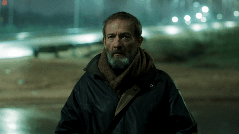 Um plano médio de César (Pablo Limarzi) no filme 'Depois, a Névoa'. Ele é um homem de meia-idade com barba e cabelo grisalhos, vestindo um casaco de couro escuro e um cachecol marrom grosso. Ele olha diretamente para a câmera com uma expressão séria, em um ambiente externo à noite, com luzes de rua ou carros desfocadas ao fundo.