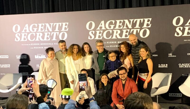 Fotografia. Elenco e equipe do filme O Agente Secreto posam juntos para fotos em frente a um painel preto com o título do filme em letras brancas, que inclui logotipos de apoiadores e patrocinadores. As pessoas sorriem e se abraçam enquanto os jornalistas fotografam a cena com celulares e câmeras.