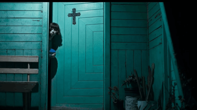 Cena do filme Delírios (2024). Uma menina de cabelos curtos e expressão assustada espreita pela fresta de uma porta verde com uma cruz de ferro pregada no alto. O ambiente é escuro e frio, com um banco de madeira e vasos de plantas secas ao lado. A iluminação destaca a textura da madeira, criando um clima de suspense e isolamento.