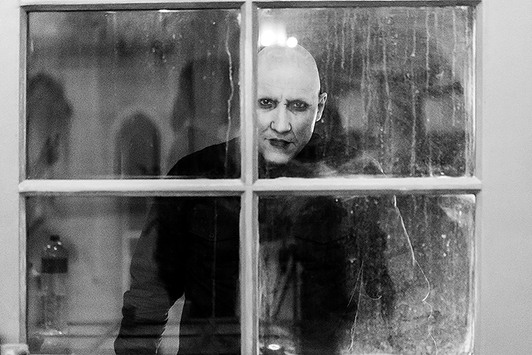 Cena do filme NosferatuNa imagem, em preto e branco, Nosferatu está de frente para a câmera, mas atrás de uma janela manchada. Ele é uma criatura de pele clara, cabeça sem cabelo, lábios escuros e olhos sombreados. Está vestindo uma roupa que cobre todo seu corpo, na cor preta. A janela é dividida de forma quadricular, em quatro partes menores. 