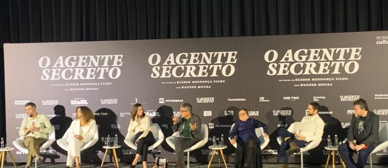 Coletiva de imprensa do filme Agente Secreto. O elenco, diretor e a produtora estão sentados em cadeiras enquanto um painel com o nome do filme está atrás. O diretor está falando no microfone