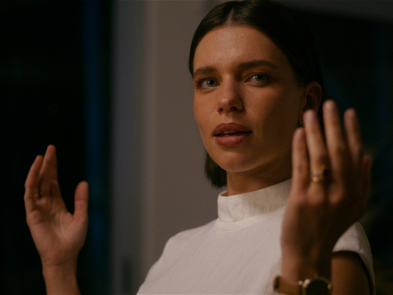 Cena do filme Virtuosas (2025). Na imagem, uma mulher está em um ambiente interno, iluminado por uma luz suave. Ela tem o cabelo curto, de um tom loiro meio escuro e veste uma blusa branca de gola alta. Seu olhar está atento e suas mãos estão erguidas