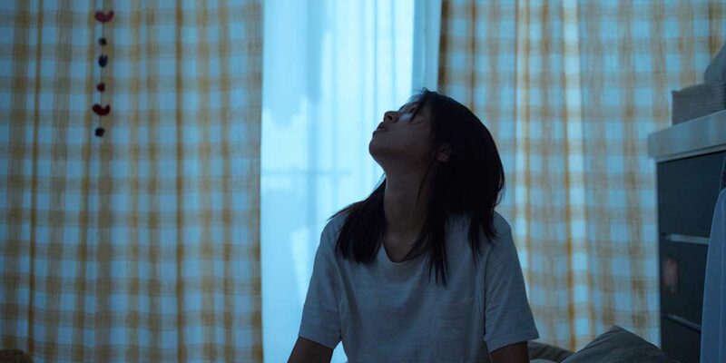 Cena do filme O Mundo do Amor. Uma jovem está sentada em sua cama, com expressão abatida, olhando para o teto. Ela veste uma camiseta branca e seu cabelo escuro cai sobre os ombros. O quarto está iluminado por uma luz azulada que entra pelas cortinas de padrão xadrez amarelo e branco, criando uma atmosfera melancólica e introspectiva.