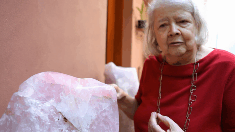 Em um plano médio, Amelia Toledo, uma mulher mais velha de cabelos brancos, está ao lado de uma grande pedra bruta de quartzo rosa. Ela veste uma blusa vermelha e um colar de corrente prateada, e olha diretamente para a câmera com uma expressão séria.