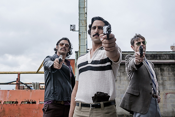 Cena do filme Homem de OuroNa imagem, três homens brancos estão no centro, de frente, apontando uma pistola. O homem do meio é o mais alto, tem cabelos escuros na altura do pescoço e usa um bigode bem grosso. Ele está de cara fechada, franzindo a testa. Veste uma camiseta gola V na cor branca, com duas faixas horizontais na cor preta no peito. A calça bege está apertada com um cinto preto, um par de luvas está enfiado na cintura. Ele segura a arma com a mão esquerda. Do seu lado direito, há um homem de cabelos curtos, lisos e loiros, que veste paletó cinza, terno e calça azul. No rosto, ele usa um óculos escuro no modelo aviador, além de um cavanhaque branco. Empunha a pistola na mão direita, onde há um anel dourado no dedo mindinho. A outra mão está apoiada na cintura. Já do lado esquerdo, está um homem com um cabelo escuro também um pouco mais longo. Ele usa bigode e óculos escuros, está vestindo uma jaqueta preta de couro e calça marrom. A arma também está em sua mão direita. O céu está nublado e o cenário é um porto, há uma embarcação laranja atrás deles, do lado esquerdo, enquanto do direito uma parede de tijolos cinzas. 