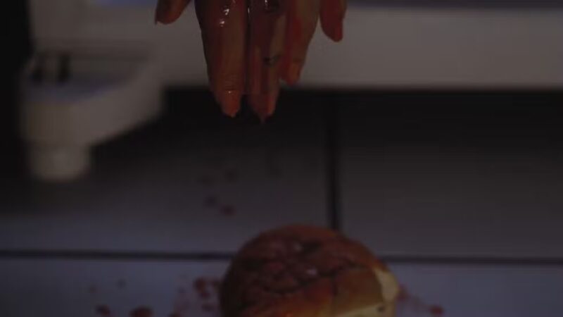 Cena do filme X-SlasherNa imagem, há um lanche no chão na parte inferior e uma mão estendida na parte superior, saindo de dentro de uma geladeira. A mão está escorrendo sangue, que cai no lanche e no chão. O eletrodoméstico é branco, assim como o piso no chão. O cenário é noturno. 