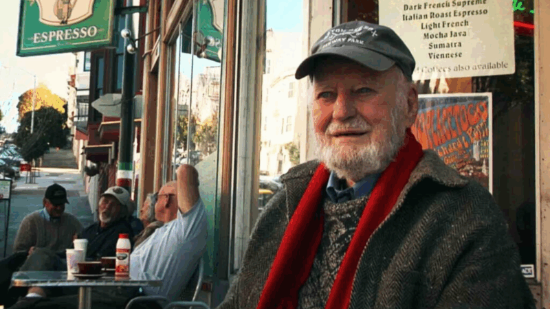 O poeta Lawrence Ferlinghetti, em cena do documentário 'A Última Batida', sentado em uma mesa na área externa de um café. Ele usa um boné escuro, um cachecol vermelho e um casaco texturizado, e sorri levemente para a câmera. Ao fundo, desfocados, outros clientes sentam-se em mesas e uma placa verde de 'ESPRESSO' se projeta da fachada.