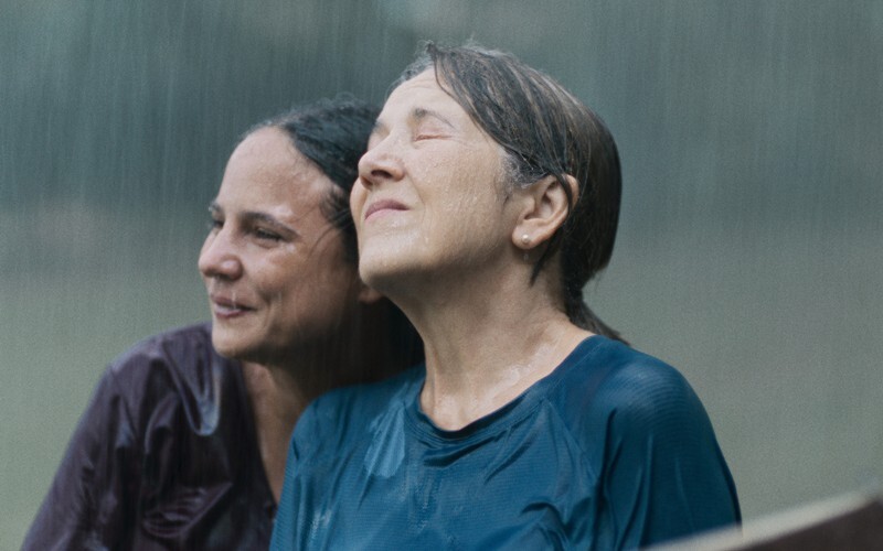Cena do filme Querido Trópico. Duas mulheres adultas estão lado a lado sob a chuva forte. A mulher da direita é branca, de cabelos grisalhos, veste uma camiseta azul e está com os olhos fechados. Ao seu lado e com o rosto colado no seu, há uma mulher de tom de pele mais escuro e cabelos castanhos e veste uma camiseta bordô. Essa está sorrindo olhando para frente. As roupas de ambas estão encharcadas e o cenário é de chuva forte.
