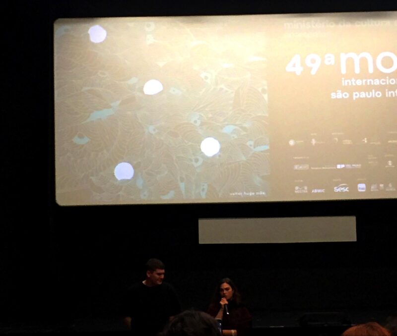 Joscha Bongard, um homem branco de cabelo escuro veste uma camisa preta. Ao seu lado, está uma mulher branca de cabelo escuro segurando um microfone. Eles estão em uma sala de cinema e ao fundo aparece a tela com a ilustração e logo da 49ª edição da Mostra.