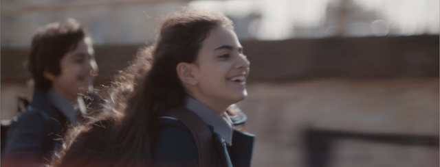 Cena do filme Um Mundo Triste e Belo (2025). Em um dia ensolarado, uma jovem de cabelos longos e soltos corre sorrindo, com o vento batendo em seu rosto. Ela veste um casaco escuro que faz parte de seu uniforme escolar e carrega uma mochila nos ombros. Ao fundo, um menino de cabelo curto também corre, parcialmente desfocado. A luz suave e o movimento da cena transmitem uma sensação de liberdade, juventude e alegria passageira.