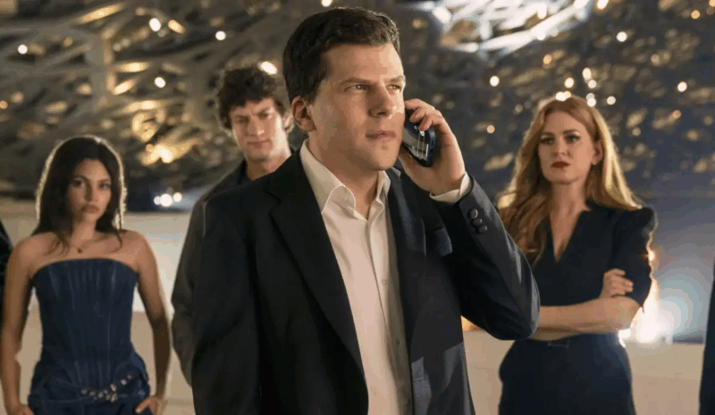 Cena de Truque de Mestre - O 3° Ato. A cena mostra um grupo em um ambiente moderno e elegante, com foco em um homem de terno escuro falando ao telefone, cercado por pessoas em trajes sofisticados. As expressões variam entre seriedade e neutralidade, sugerindo tensão. A iluminação suave e o design contemporâneo do espaço criam um clima cinematográfico, com tons neutros e detalhes controlados que reforçam um ar de suspense.