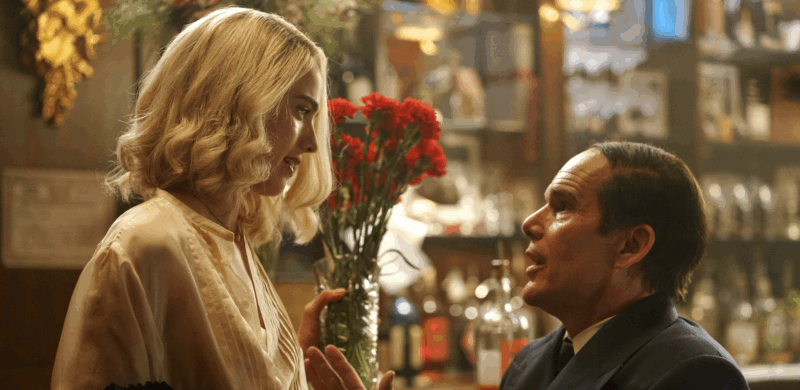  Cena do filme Blue Moon. Na imagem, há uma mulher branca de cabelos louros e blusa bege. Ela olha para um homem branco de cabelos castanhos com terno. Eles estão em um bar. 