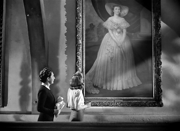 Cena de Rebecca, A Mulher Inesquecível. Nesta imagem em preto e branco, duas mulheres estão de costas e de lado, em um grande salão ou galeria. Elas olham para cima, admirando um retrato a óleo muito grande, de corpo inteiro, de uma mulher em um vestido de gala e chapéu, emoldurado de forma ornamentada. A mulher à esquerda na imagem usa um traje escuro e formal, com o cabelo preso. A mulher à direita usa roupas mais claras, com o cabelo solto até os ombros. A cena é capturada de um ângulo médio, destacando a imponência do retrato em relação às duas espectadoras.