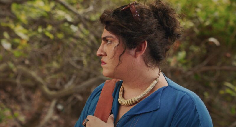 Cena do filme Morte e Vida Madalena Na imagem, Madalena está de perfil, olhando para o lado esquerdo. Ela veste uma camisa azul marinho, uma bolsa marrom com a alça apoiada no ombro direito e um colar grosso com discos enfileirados que parece ser feito do material sisal. Na mão esquerda, que segura a bolsa, há um anel com uma pedra vermelha no dedo do meio. Ao fundo, árvores e galhos. Madalena é travesti e possui cabelos cacheados na cor castanha, que estão presos, há um óculos de sol apoiado em sua cabeça. 