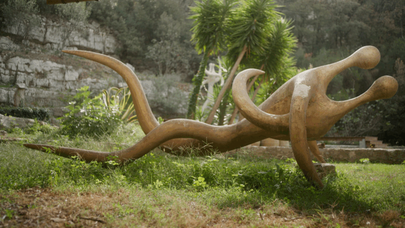 Uma escultura alta e abstrata de Robert Roussil, de cor clara, com uma forma humanóide estilizada. A figura, que se assemelha a uma pessoa dançando com braços curvos e erguidos, está posicionada ao ar livre contra um fundo escuro de um paredão rochoso.