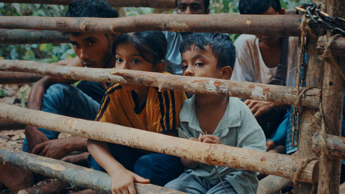 Cena do filme Terra Perdida. Somira, vestindo uma camiseta laranja e calça azul, e Shafi, com uma camiseta clara e calça cinza, observam o exterior por entre as frestas de uma cerca de madeira, ao lado de outros refugiados