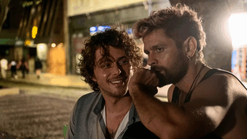 Uma cena noturna de Ruas da Glória que mostra os personagens Gabriel (Caio Macedo) à esquerda e Adriano (Alejandro Claveaux) à direita, sentados próximos em um ambiente urbano. Gabriel, de camisa clara, sorri e olha para Adriano. Adriano, que tem barba e cabelo mais curto com pontas rosas, olha intensamente para Gabriel com uma expressão séria, o punho apoiado perto da boca. A iluminação quente e suave destaca a conexão e a intimidade entre os dois.