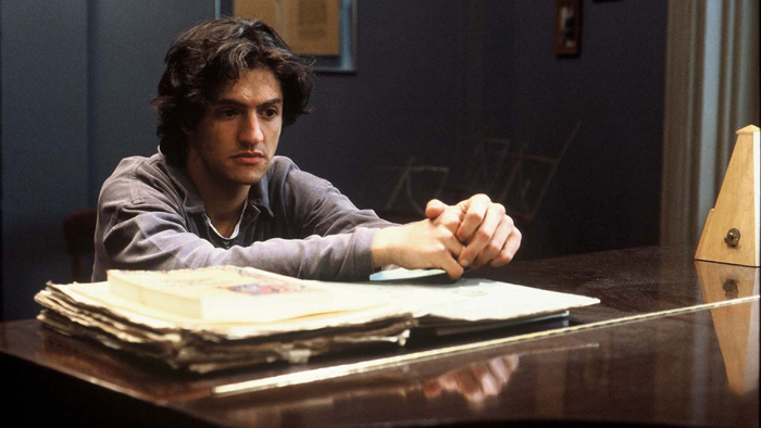 Cena do filme Tônica Dominante. Um jovem de cabelos escuros está sentado à frente de um piano, com as mãos cruzadas e expressão introspectiva. Em cima do instrumento há partituras e livros empilhados. Ao fundo há uma parede azul com dois quadros pendurados e 3 porta-partituras.