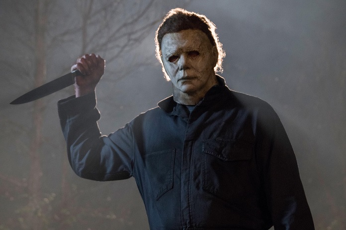 Cena de Halloween de 2018. O personagem de terror Michael Myers em um cenário escuro e enevoado. Ele usa sua icônica máscara branca e um macacão azul escuro, e segura uma grande faca de cozinha levantada na mão direita, olhando fixamente para a frente.