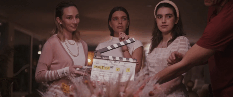 Cena dos bastidores do filme Virtuosas (2025). Três mulheres estão posicionadas lado a lado, enquanto uma claquete é segurada à frente delas. A mulher à esquerda veste um cardigã rosa e um colar de pérolas; a do centro, uma blusa branca de gola alta; e a da direita, um vestido rosa claro com uma tiara. Elas olham concentradas para alguém fora do quadro, em um ambiente elegante e bem iluminado, sugerindo um momento de preparação para a gravação