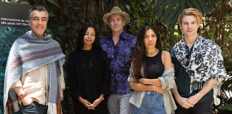 Foto posada, em plano médio, de cinco pessoas em pé, lado a lado, em um ambiente externo com uma parede de folhagens densas ao fundo. Da esquerda para a direita estão: um homem usando seu xale geométrico cinza e azul; uma mulher negra com longas tranças e blusa preta de mangas compridas; o crítico Peter Debruge ao centro, usando chapéu e camisa roxa com estampa floral; uma mulher de cabelos longos e escuros vestindo regata preta e segurando uma camisa jeans; e um homem loiro vestindo um lenço grande preto e branco sobre uma camisa escura. À esquerda, vê-se parte do painel preto com a identidade visual da 49ª Mostra.