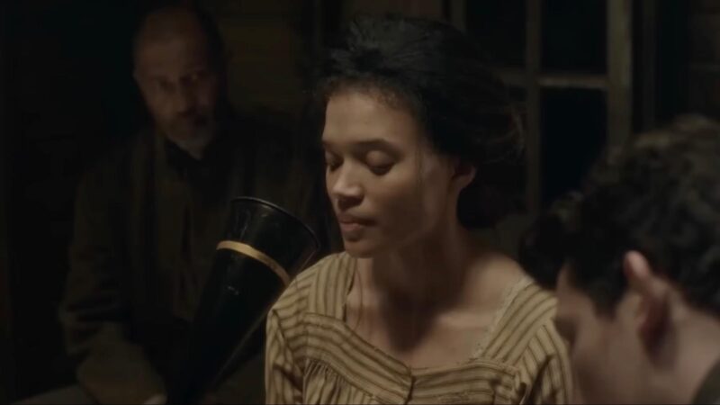 Uma mulher negra de cabelos escuros e vestido bege listrado está sentada cantando em frente ao fonógrafo. Ao fundo, aparece um homem olhando a moça cantar.