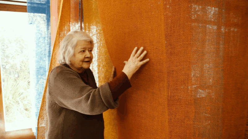 Amelia Toledo, uma mulher mais velha de cabelos brancos, é vista de perfil, tocando com a mão direita uma grande instalação têxtil laranja, semelhante a uma malha. Ela veste um suéter marrom e olha para a obra com um leve sorriso, em um ambiente interno com uma janela ao fundo.