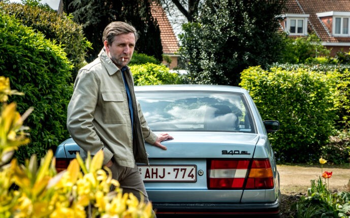 Cena do filme Maturidade. Na imagem,é possível ver um homem branco de cabelo castanho, vestindo uma camisa azul, um casaco bege e calças cinzas. Na sua boca, há um cigarro aceso. Ele está com a mão esquerda encostada no porta-malas de um carro na cor azul-claro. Ao fundo da imagem, é possível ver aspectos de uma vizinhança residencial, com os telhados das casas em marrom-alaranjado e arbustos grandes com folhas verdes. 