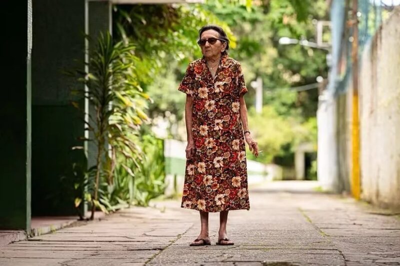 Cena do filme O Agente Secreto. Dona Sebastiana, uma mulher idosa de pele branca e cabelos castanhos caminha sozinha em uma calçada arborizada. Ela usa um vestido estampado com flores em tons de vermelho, marrom e bege, chinelos e óculos escuros. Na mão esquerda, ela segura um cigarro.