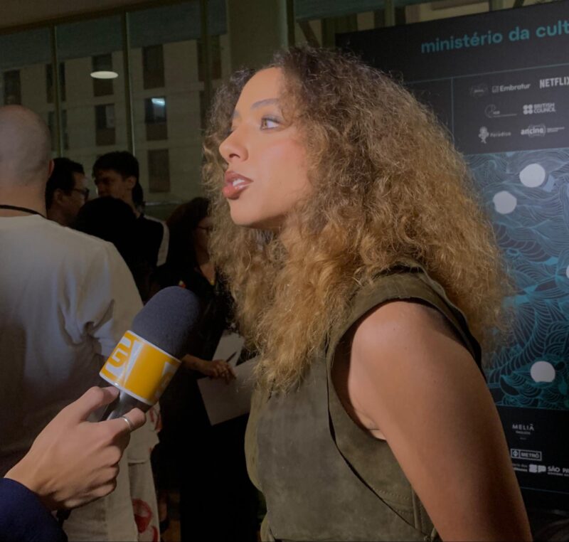 Tapete vermelho do filme Agente Secreto. Uma mulher, com cabelos cacheados e com um vestido verde está falando ao microfone. O microfone está sendo segurado por uma mão de um homem branco. Ao fundo, está o painel da 49ª Mostra de Cinema em São Paulo com várias pessoas ao redor