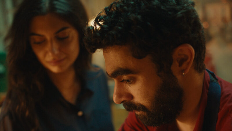 Cena do filme Um Mundo Triste e Belo (2025). Em um ambiente interno com luz amarelada e suave, o homem está em primeiro plano, olhando para baixo de forma pensativa. Ele tem barba escura e usa uma camisa vermelha. Atrás dele, a mulher o observa com um olhar terno, vestindo uma blusa azul. A cena transmite intimidade, reflexão e afeto contido