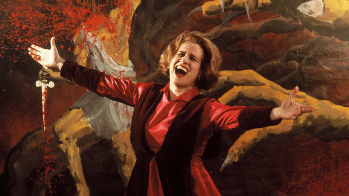 Cena do filme Tônica Dominante. Uma mulher vestindo um vestido vermelho está de braços abertos em gesto expansivo diante de uma pintura expressionista com tons de vermelho e amarelo fortes. Ela está com a boca aberta, gritando com intensidade.