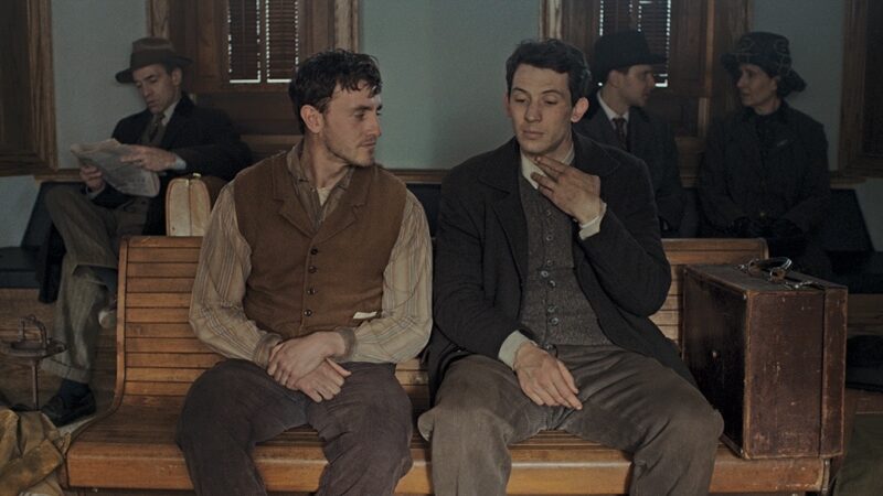 Cena do filme A História do Som: Dois homens, Lionel e David ( da esquerda para a direita), brancos e de cabelos castanhos, estão sentados lado a lado em um banco de madeira em uma sala de uma estação de trem antiga. Ambos vestem roupas de época, em tons marrons. Lionel olha para o outro enquanto conversa; David toca o próprio pescoço com a mão e parece pensativo. Ao fundo, há outras pessoas sentadas; um homem lê o jornal e um casal conversa entre si.