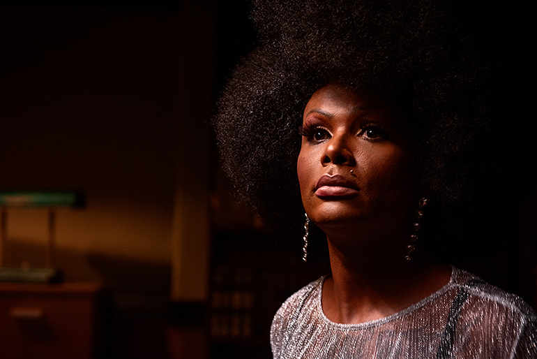 Em uma cena de Ruas da Glória, a personagem Mônica, interpretada por Diva Menner, é vista em um close-up de perfil. Ela é uma mulher negra com um cabelo afro volumoso, maquiagem marcante, um piercing no lábio e brincos longos. Mônica veste uma blusa prateada brilhante e olha para cima, com o rosto parcialmente iluminado por uma luz quente que vem da direita, deixando a outra metade em sombra profunda e criando um clima pensativo e dramático.