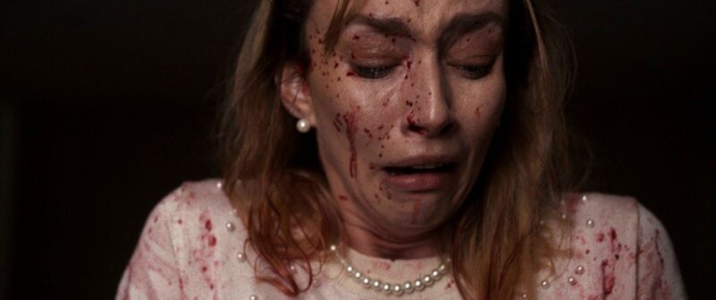 Cena do filme Virtuosas. Close-up de uma mulher loira com o rosto coberto de sangue, chorando e com os olhos fechados. Ela usa um colar de pérolas e uma blusa branca com detalhes em pérolas, também manchada de sangue. Há uma luz suave iluminando seu rosto contra um fundo escuro.