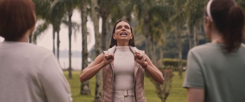 Cena do filme Virtuosas. Uma mulher com cabelo castanho escuro e liso, vestindo um colete bege sobre uma blusa de gola alta da mesma cor e calça bege, está de frente para a câmera com a boca aberta e os braços levemente erguidos, com os punhos cerrados. Ela está ao ar livre, em meio a palmeiras, e outras duas mulheres de costas para a câmera aparecem nas laterais da imagem.